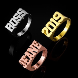 Op de afbeelding: Drie gepersonaliseerde ringen in zilver, goud en roségoud. De zilveren ring spelt "ROSS" met kleine diamantaccenten. De gouden ring toont het nummer "2019". De roségouden ring spelt "JEANE". Alle ringen hebben een blokletterontwerp.