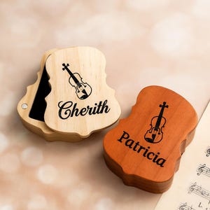 May include: Two wooden violin-shaped boxes. One is light-coloured with "Cherith" and a violin illustration, the other is brown with "Patricia" and a violin illustration. The boxes are open, revealing a black interior.