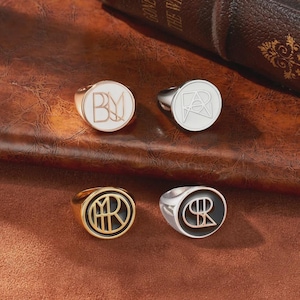 May include: Four signet rings with different designs. Two rings have gold bands, one with a white face and the other with a black face. The other two rings have silver bands, one with a white face and the other with a silver face. Each ring has a unique monogram.