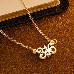 May include: A gold-colored necklace with a delicate chain. The pendant features the number "316" in an elegant, stylized font. The necklace is displayed on a brown surface, possibly leather or a book.