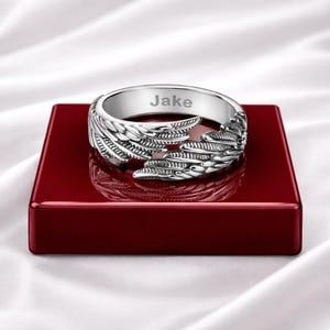 Puede incluir: Anillo plateado con dise&ntilde;o de alas y el nombre "Jake" grabado. El anillo se muestra sobre una superficie rectangular roja.