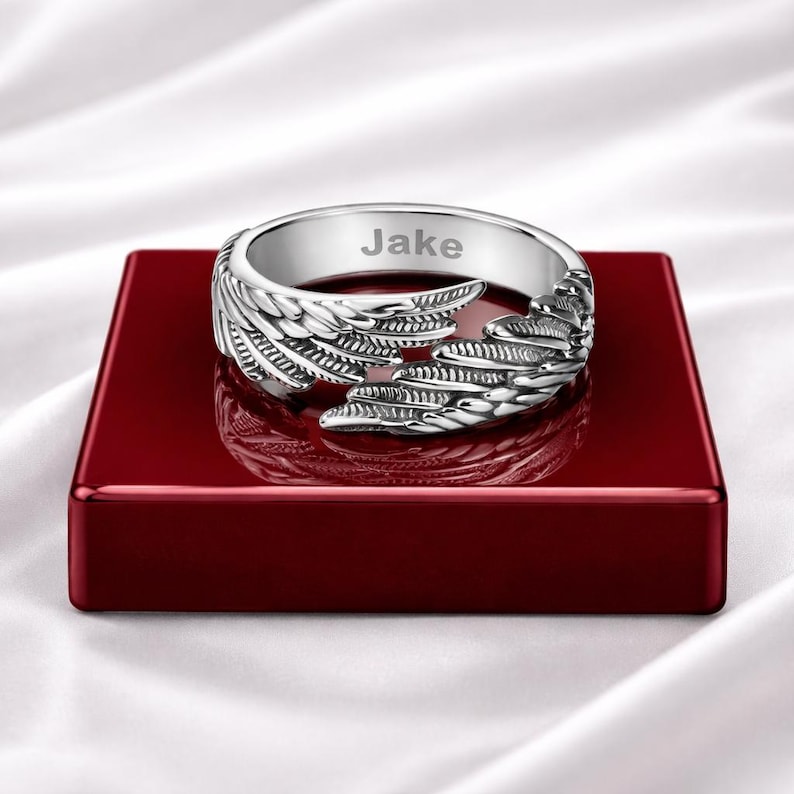 Puede incluir: Anillo plateado con dise&ntilde;o de alas y el nombre "Jake" grabado. El anillo se muestra sobre una superficie rectangular roja.