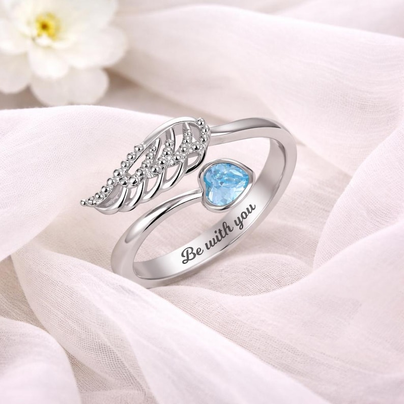Puede incluir: Anillo plateado con una piedra preciosa azul en forma de coraz&oacute;n y un dise&ntilde;o de ala. El anillo tiene la inscripci&oacute;n "Be with you". El anillo est&aacute; sobre un fondo de tela blanca y suave.