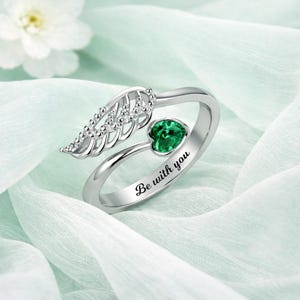 Puede incluir: Anillo plateado con dise&ntilde;o de ala y gema verde en forma de coraz&oacute;n. El anillo tiene la inscripci&oacute;n "Be with you". Fondo de tela azul claro y suave.