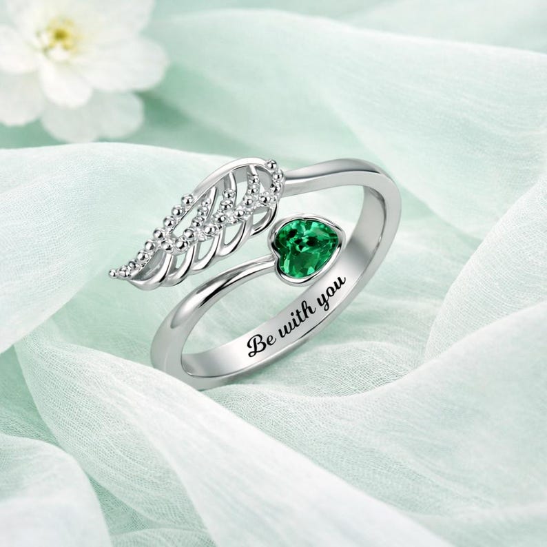 Puede incluir: Anillo plateado con dise&ntilde;o de ala y gema verde en forma de coraz&oacute;n. El anillo tiene la inscripci&oacute;n "Be with you". Fondo de tela azul claro y suave.