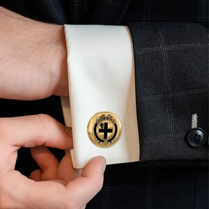 K&ouml;nnte beinhalten: Goldfarbene Manschettenkn&ouml;pfe mit einem schwarz-wei&szlig;en Emblem. Die runden Manschettenkn&ouml;pfe tragen den Text "HOUSE OF BURKE". Das Emblem enth&auml;lt ein Kreuz und einen L&ouml;wen. Die Manschettenkn&ouml;pfe befinden sich an einem wei&szlig;en Hemd&auml;rmel.