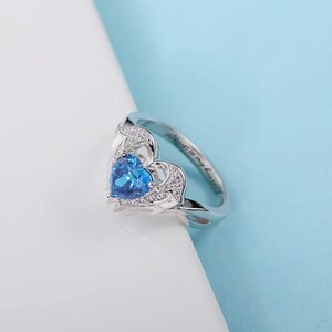 May include: A silver ring with a heart-shaped blue gemstone at the center. The ring has a delicate design with small, clear stones around the heart. The inscription "Always in my heart" is engraved on the band.