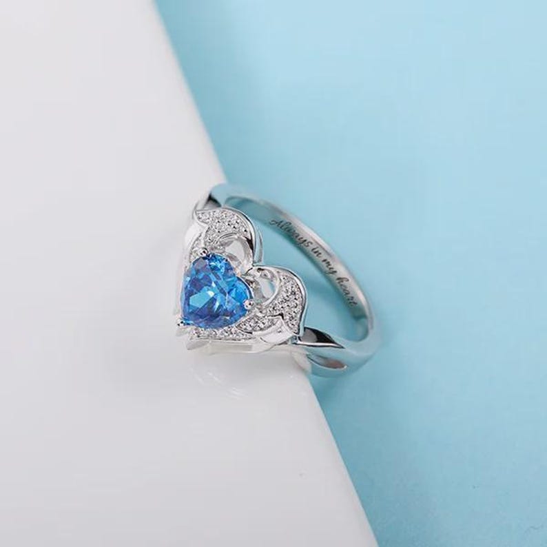 May include: A silver ring with a heart-shaped blue gemstone at the center. The ring has a delicate design with small, clear stones around the heart. The inscription "Always in my heart" is engraved on the band.