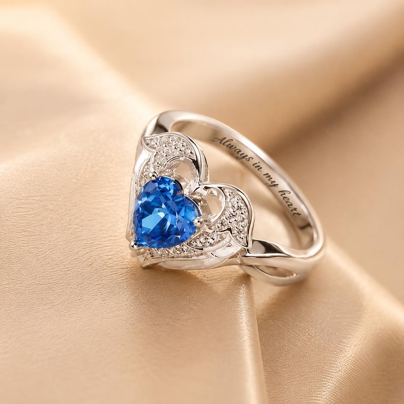 May include: A silver ring with a heart-shaped blue gemstone at the center. The ring features a delicate design with small, clear accent stones and the inscription "Always in my heart". The ring is set against a soft, golden background.