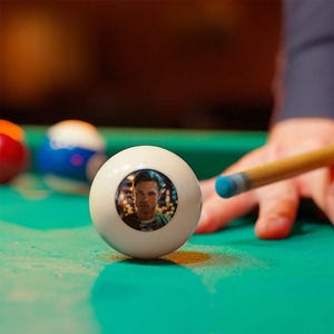Puede incluir: Una bola de billar blanca con un retrato, colocada sobre una mesa de billar verde. Un taco de billar con una punta azul apunta a la bola. Otras bolas de billar de colores están en el fondo.