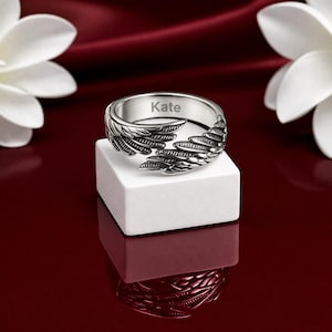Puede incluir: Anillo plateado con dise&ntilde;o de alas, grabado con el nombre "Kate". Se muestra sobre un bloque blanco, sobre un fondo rojo oscuro, con flores blancas al fondo. El dise&ntilde;o del anillo es detallado.