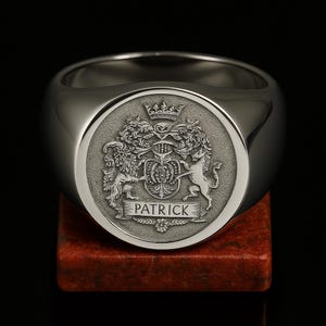 May include: A silver signet ring with a detailed crest design. The crest features a crown, shield, and two heraldic animals, with the name "PATRICK" inscribed below. The ring is displayed on a red surface.