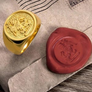 May include: A gold signet ring featuring a raised relief of a lion holding a sword on its circular face. Beside the ring is a round, dark red wax seal stamp that displays an identical embossed lion and sword design on its surface.