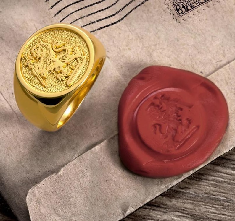May include: A gold signet ring featuring a raised relief of a lion holding a sword on its circular face. Beside the ring is a round, dark red wax seal stamp that displays an identical embossed lion and sword design on its surface.