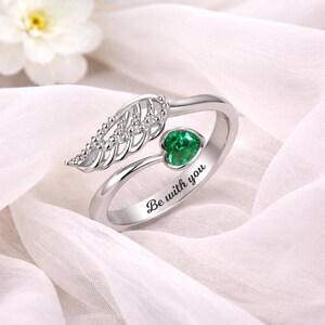 Puede incluir: Anillo plateado con una gema verde en forma de coraz&oacute;n y un dise&ntilde;o de ala. El anillo tiene la inscripci&oacute;n "Be with you". El anillo est&aacute; sobre un fondo blanco.