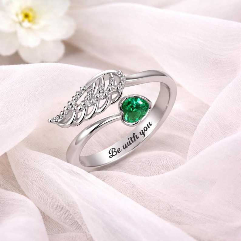 Puede incluir: Anillo plateado con una gema verde en forma de coraz&oacute;n y un dise&ntilde;o de ala. El anillo tiene la inscripci&oacute;n "Be with you". El anillo est&aacute; sobre un fondo blanco.