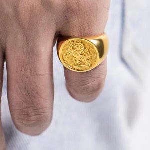 May include: A gold-toned signet ring featuring a circular face with an embossed design of a figure on horseback wielding a sword. The ring has a smooth, polished band and a textured, recessed background within the circular setting.