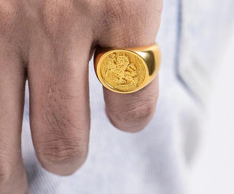 May include: A gold-toned signet ring featuring a circular face with an embossed design of a figure on horseback wielding a sword. The ring has a smooth, polished band and a textured, recessed background within the circular setting.