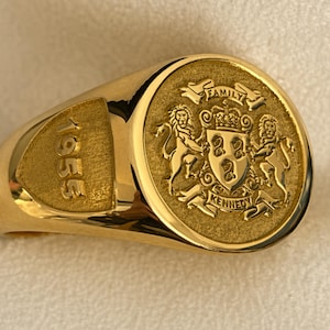 Può includere: Un anello con sigillo d'oro con un stemma dettagliato. Lo stemma presenta due leoni, uno scudo e le parole "FAMILY" e "KENNEDY". L'anno "1955" è inciso sul lato. Un gioiello classico.