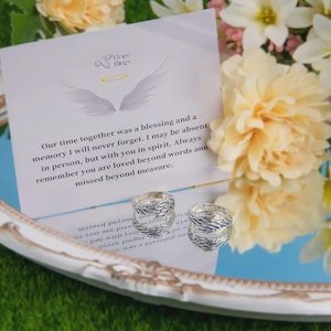 Puede incluir: Dos anillos de plata con dise&ntilde;o de plumas se exhiben sobre una superficie reflectante, junto a una tarjeta con el texto "Hope Rings" y un mensaje de recuerdo. Los anillos se colocan sobre un fondo de flores.