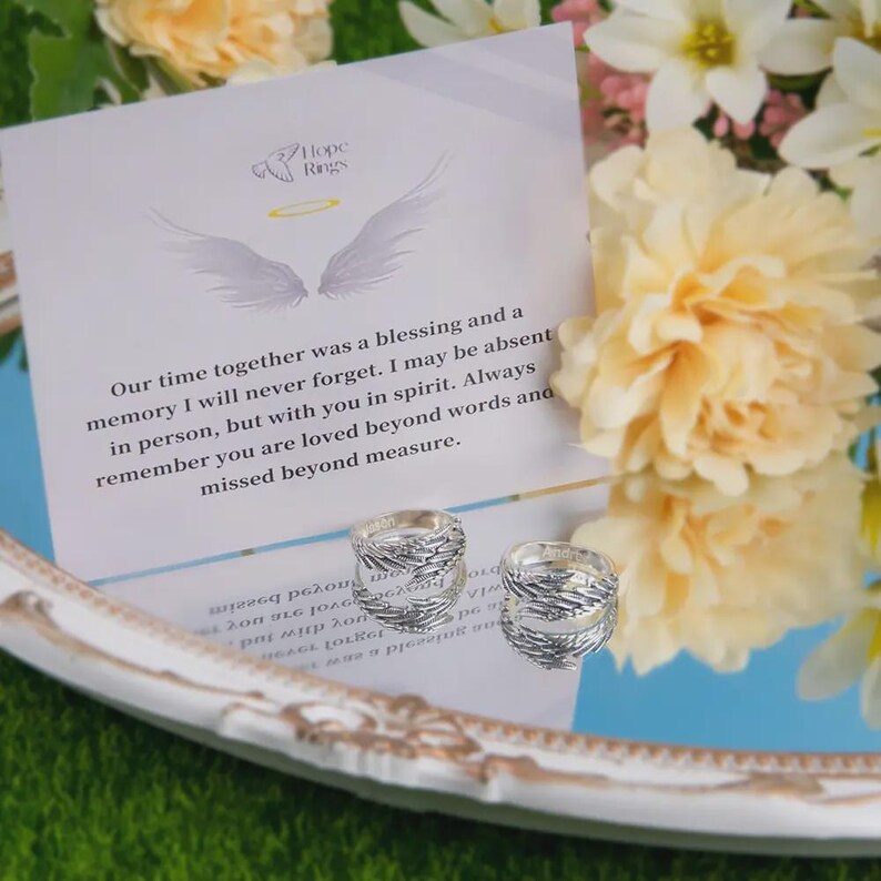 Puede incluir: Dos anillos de plata con dise&ntilde;o de plumas se exhiben sobre una superficie reflectante, junto a una tarjeta con el texto "Hope Rings" y un mensaje de recuerdo. Los anillos se colocan sobre un fondo de flores.