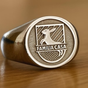 May include: A silver-toned signet ring featuring an oval face with an engraved crest. The crest displays a rabbit silhouette above the text FAMILIA CASA, set against a background of diagonal lines and a dotted pattern at the base.