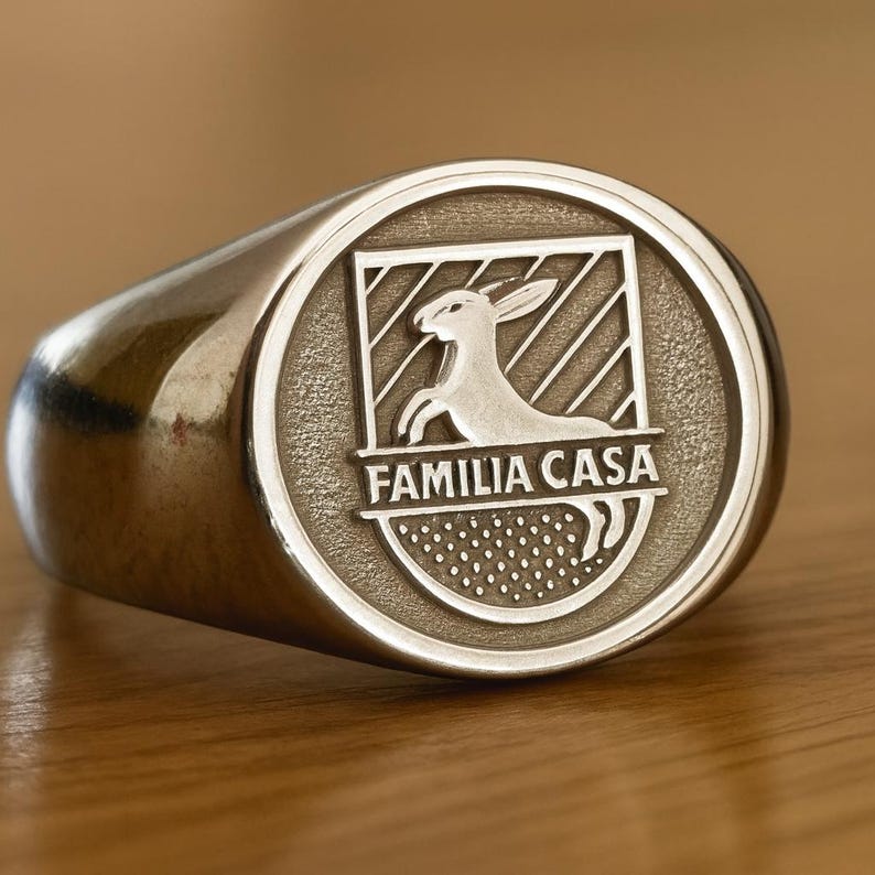 May include: A silver-toned signet ring featuring an oval face with an engraved crest. The crest displays a rabbit silhouette above the text FAMILIA CASA, set against a background of diagonal lines and a dotted pattern at the base.