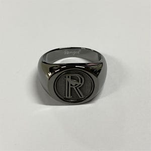 May include: A polished gunmetal signet ring featuring a circular face with a large engraved letter R and a small bird silhouette. The interior band is engraved with the cursive text Rangel in a matching metallic finish.