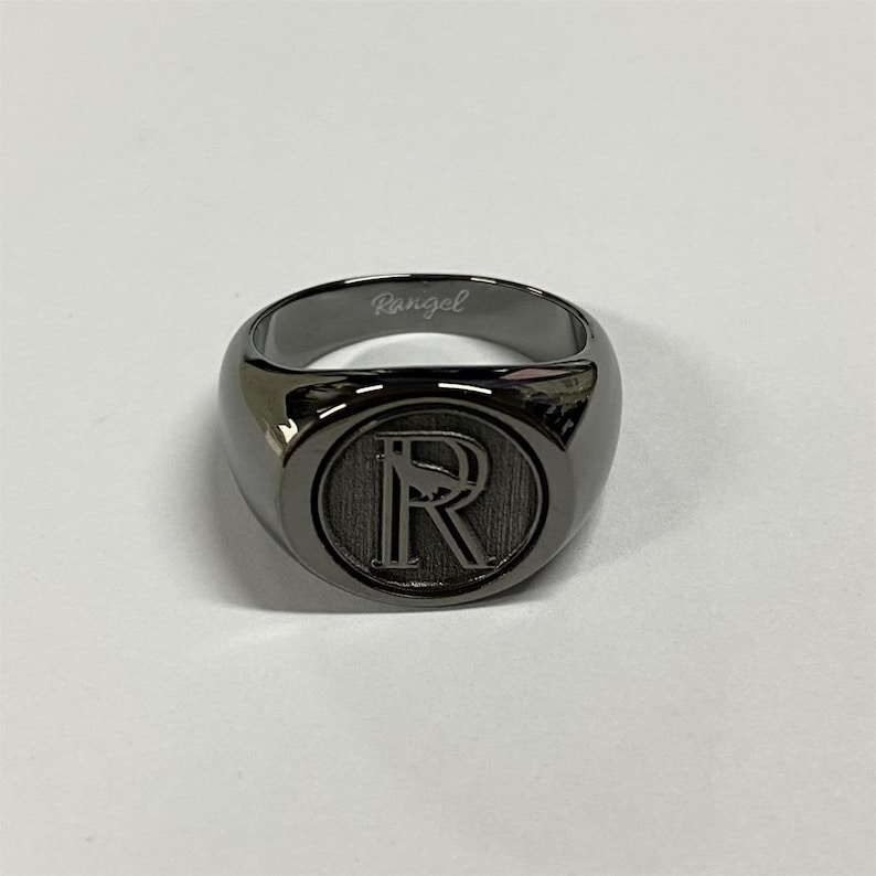 May include: A polished gunmetal signet ring featuring a circular face with a large engraved letter R and a small bird silhouette. The interior band is engraved with the cursive text Rangel in a matching metallic finish.
