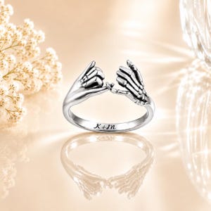 Puede incluir: Un anillo plateado con dos manos en un gesto de promesa. Una mano es lisa, la otra esquelética. El anillo está grabado con las iniciales "K+m". El anillo se muestra sobre una superficie reflectante.