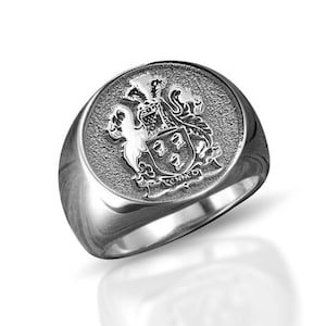 May include: A silver-toned signet ring featuring a circular face with a textured background. The center displays an engraved heraldic crest with a shield and the name KENNEDY inscribed on a banner at the base of the emblem.