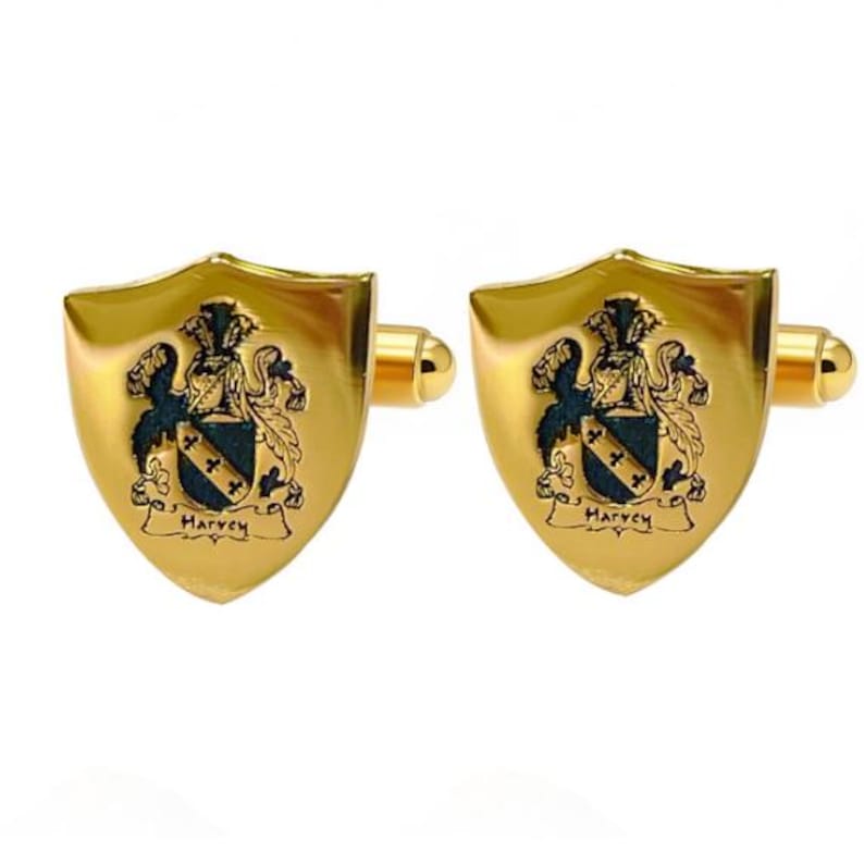 K&ouml;nnte beinhalten: Ein Paar goldfarbene Manschettenkn&ouml;pfe mit Schilddesign. Jeder Manschettenknopf zeigt ein Wappen mit einem blau-schwarzen Emblem und dem Schriftzug "Harvey". Die Manschettenkn&ouml;pfe haben eine polierte Oberfl&auml;che.