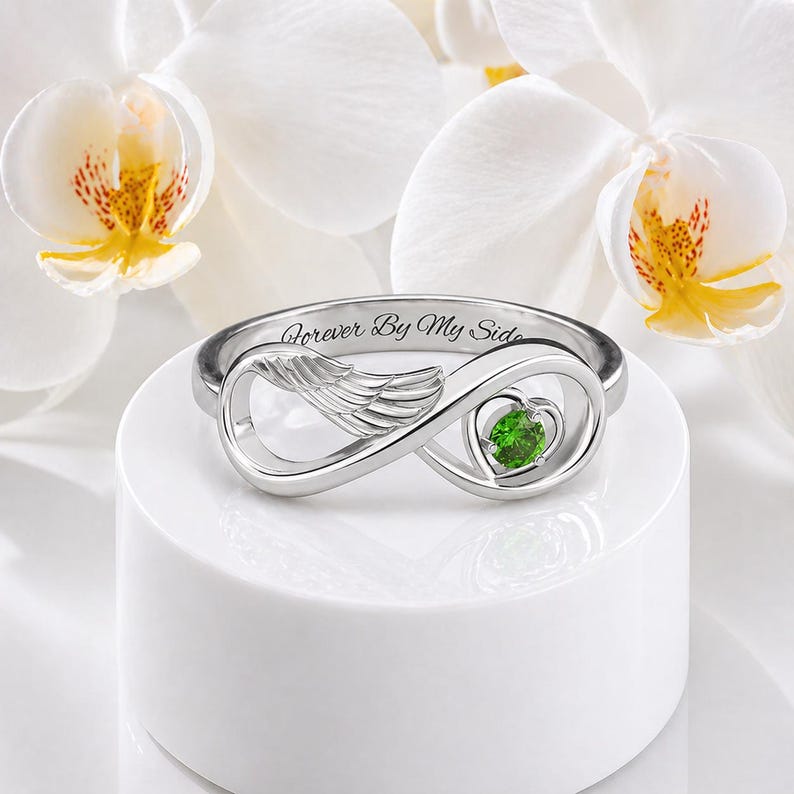May include: A silver ring with an infinity symbol design, featuring a wing detail and a heart-shaped green gemstone. The ring is engraved with the words "Forever By My Side". The ring is displayed on a white pedestal, with white orchids in the background.