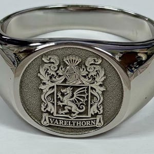 May include: A polished silver-tone signet ring featuring a circular face with an engraved coat of arms. The design includes a central dragon, a helmet crest, ornate scrollwork, and the text VARELTHORN inscribed on a banner at the base.