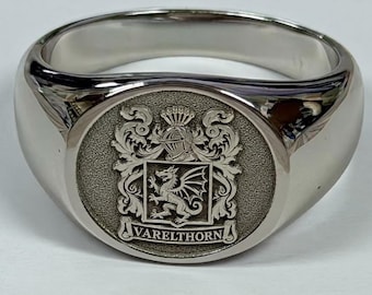 Peut inclure: Une bague chevalière en métal argenté poli avec un plateau circulaire gravé d'armoiries. Le motif comprend un dragon central, un heaume, des volutes ornementales et le texte VARELTHORN inscrit sur une bannière à la base.