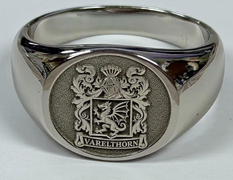 May include: A polished silver-tone signet ring featuring a circular face with an engraved coat of arms. The design includes a central dragon, a helmet crest, ornate scrollwork, and the text VARELTHORN inscribed on a banner at the base.