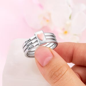Può includere: Un anello d'argento con tre fasce, ognuna incisa con un nome: Prad, Jacob e tmily. Un elemento rettangolare con un cuore rosa è centrato sull'anello. L'anello è tenuto in mano su uno sfondo rosa tenue.