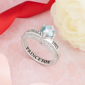 May include: A silver ring featuring a large, light blue gemstone. The ring is engraved with the name "ALTHAEA" and "PRINCETON" and the year "2025". The band is encrusted with small, clear gemstones. The background includes a red ribbon and a white flower.