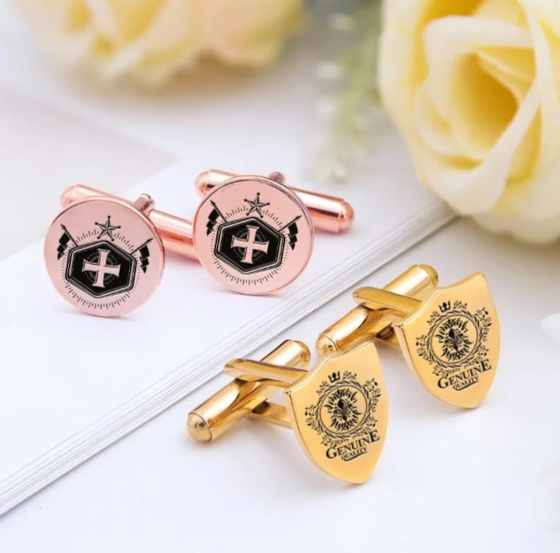 K&ouml;nnte beinhalten: Manschettenkn&ouml;pfe in Ros&eacute;gold und Gold. Die Manschettenkn&ouml;pfe in Ros&eacute;gold haben ein kreisf&ouml;rmiges Design mit einem Kreuz- und Stern-Emblem. Die goldenen Manschettenkn&ouml;pfe sind schildf&ouml;rmig mit einem floralen Design und dem Schriftzug "GENUINE QUALITY".