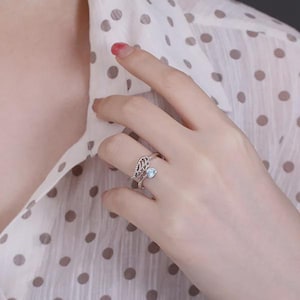 Puede incluir: Un anillo plateado con un delicado dise&ntilde;o de ala y una peque&ntilde;a gema azul claro. El anillo se lleva en un dedo, mostrando sus intrincados detalles y su elegante estilo. El dise&ntilde;o del anillo recuerda a un ala de &aacute;ngel.