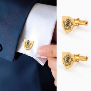 K&ouml;nnte beinhalten: Goldfarbene manschettenkn&ouml;pfe in schildform mit einem dekorativen Emblem und dem Text "GENUINE QUALITY". Ein Manschettenknopf ist an einem wei&szlig;en Hemd&auml;rmel befestigt, und zwei weitere Manschettenkn&ouml;pfe werden separat gezeigt.