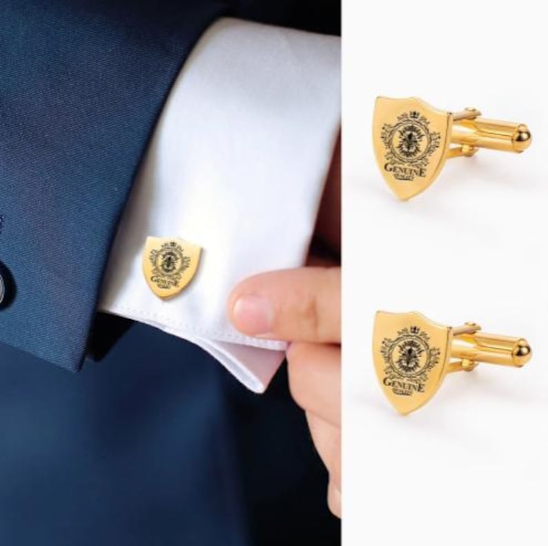 K&ouml;nnte beinhalten: Goldfarbene manschettenkn&ouml;pfe in schildform mit einem dekorativen Emblem und dem Text "GENUINE QUALITY". Ein Manschettenknopf ist an einem wei&szlig;en Hemd&auml;rmel befestigt, und zwei weitere Manschettenkn&ouml;pfe werden separat gezeigt.