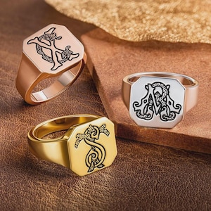 May include: Three signet rings with engraved designs. One is rose gold with a black design, another is silver with a black design, and the third is gold with a black design. The rings are displayed on a brown leather surface.