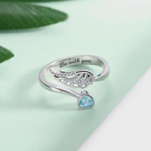 Puede incluir: Anillo plateado con dise&ntilde;o abierto, con detalle de ala de &aacute;ngel y una gema azul claro. El anillo est&aacute; grabado con las palabras "Be with you". El anillo est&aacute; sobre un fondo verde claro.