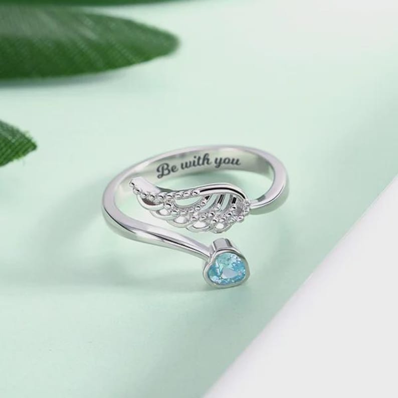 Puede incluir: Anillo plateado con dise&ntilde;o abierto, con detalle de ala de &aacute;ngel y una gema azul claro. El anillo est&aacute; grabado con las palabras "Be with you". El anillo est&aacute; sobre un fondo verde claro.