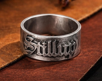 May include: A silver-colored ring with the word "Still" engraved in a gothic font. The ring has a wide band and a slightly distressed finish, set against a brown leather surface.