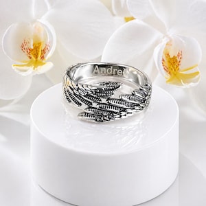 Puede incluir: Un anillo plateado con dise&ntilde;o de plumas, grabado con el nombre "Andre". El anillo se exhibe sobre un pedestal blanco, con flores de orqu&iacute;deas blancas en el fondo. El anillo tiene detalles intrincados.