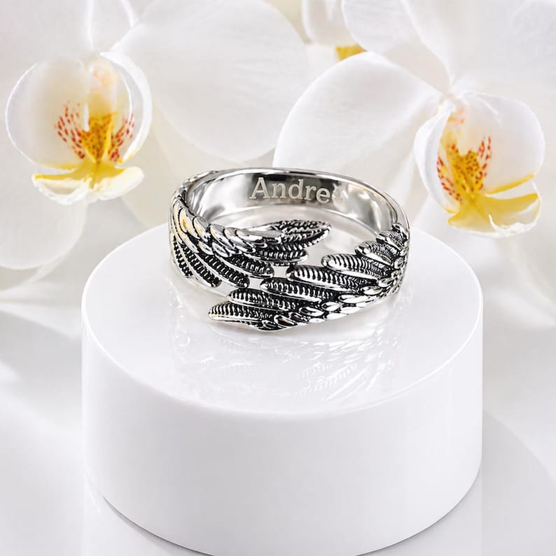 Puede incluir: Un anillo plateado con dise&ntilde;o de plumas, grabado con el nombre "Andre". El anillo se exhibe sobre un pedestal blanco, con flores de orqu&iacute;deas blancas en el fondo. El anillo tiene detalles intrincados.