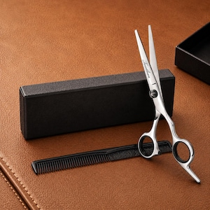 May include: A set of professional barber shears with the word "Feather" engraved on the blade, a black comb, and a black box. The shears are silver and have finger holes for easy handling. The comb has fine teeth.