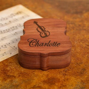 May include: A wooden box shaped like a violin, with the name "Charlotte" engraved on the front. The box has a violin illustration on the top. The box is brown and sits on a brown surface with sheet music in the background.